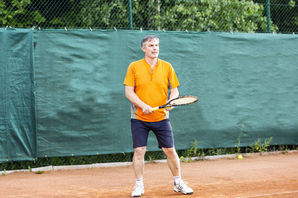 A middle-aged man plays tennis on a court with a natural earth surface on a sunny summer day. - Foto, afbeelding