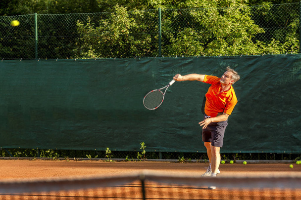 A middle-aged man plays tennis on a court with a natural earth surface on a sunny summer day. - Foto, afbeelding