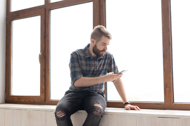 Mignon jeune homme hipster avec des vêtements élégants est assis sur les réseaux sociaux en utilisant un smartphone et Internet sans fil près de grandes fenêtres. Concept de dépendance à Internet
. - Photo, image