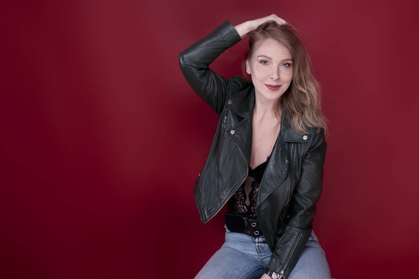 The blond girl is sitting on a high black chair in the studio, behind her is a red background. The girl was wearing jeans and a leather jacket, her hands leaned on the chair. - Photo, Image