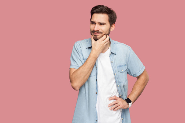 confused handsome bearded young man in blue casual style shirt scratching beard and thinking on pink background - Foto, Imagen