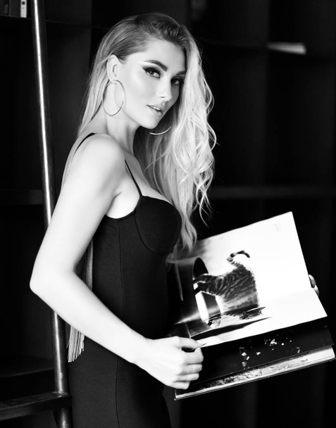 Jonge mooie vrouw in sexy kleine zwarte jurk lezen tijdschrift of boek in moderne kamer - Foto, afbeelding