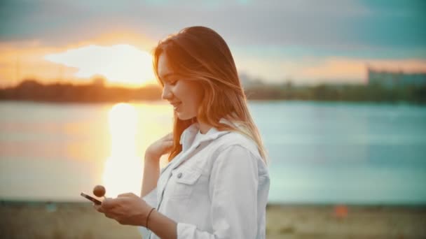 Ένα χαμογελαστή έφηβο κορίτσι στέκεται στην προκυμαία-κοιτάζοντας το τηλέφωνο-κρατώντας ένα γλειφιτζούρι-ηλιοβασίλεμα - Πλάνα, βίντεο