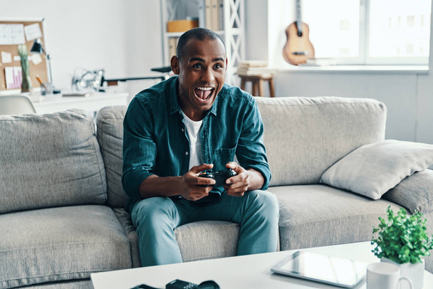 Zoveel plezier! Knappe jonge Afrikaanse man spelen video game en glimlachend zittend binnen zitten - Foto, afbeelding