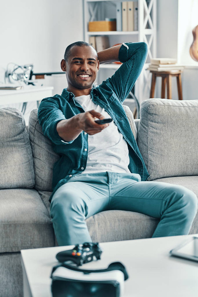 Просто расслабляюсь. Красивый молодой африканский мужчина держит телевизор и улыбается, сидя в помещении на диване
 - Фото, изображение