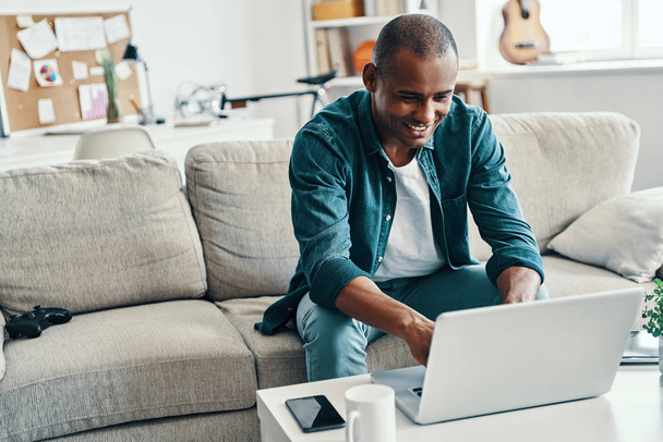 Social networking, Handsome young African man using laptop and smiling while sitting on sofa in room  - Фото, зображення