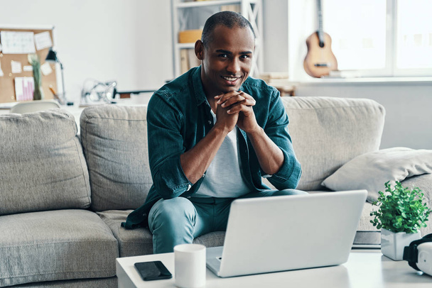 Happy and carefree. Handsome young African man using laptop and smiling while sitting indoors - Фото, изображение