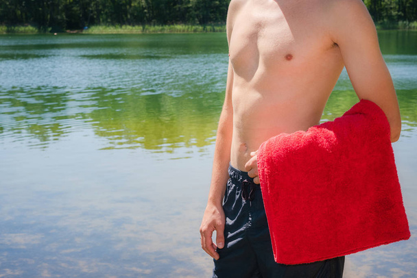 Молодой и здоровый, спортивный мужчина, стоящий на берегу озера без рубашки, держа в руках красное полотенце. Плавание, летние развлечения на природе
 - Фото, изображение