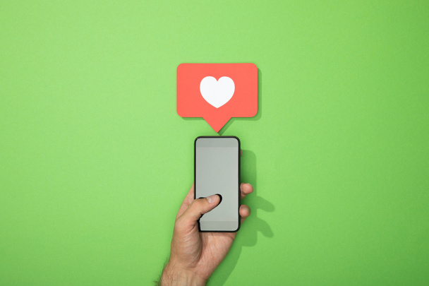 vista recortada del hombre sosteniendo el teléfono inteligente con el corazón blanco en el icono por encima de gadget en verde
 - Foto, Imagen