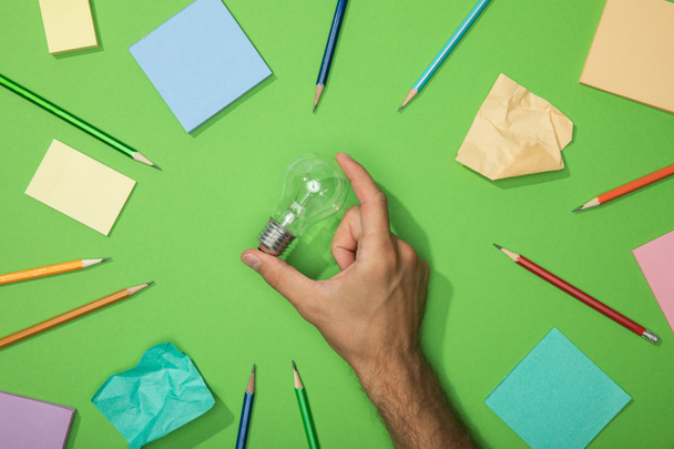 vista recortada del hombre sosteniendo la bombilla cerca de lápices dispersos y papel arrugado en verde
 - Foto, Imagen