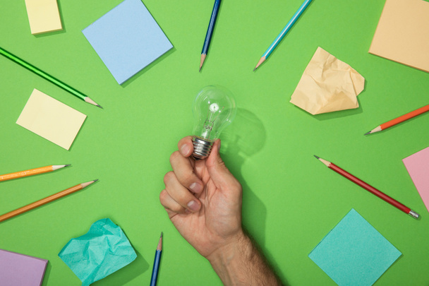 cropped view of man holding light bulb near pencils and paper on green - Photo, Image