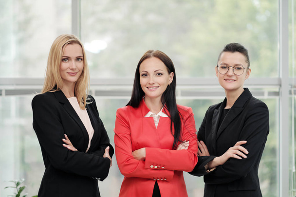 Tres jóvenes mujeres atractivas en trajes de negocios posando en el contexto de una oficina ligera. Cabeza y subordinados. Equipo de trabajo de profesionales y colegas
. - Foto, Imagen