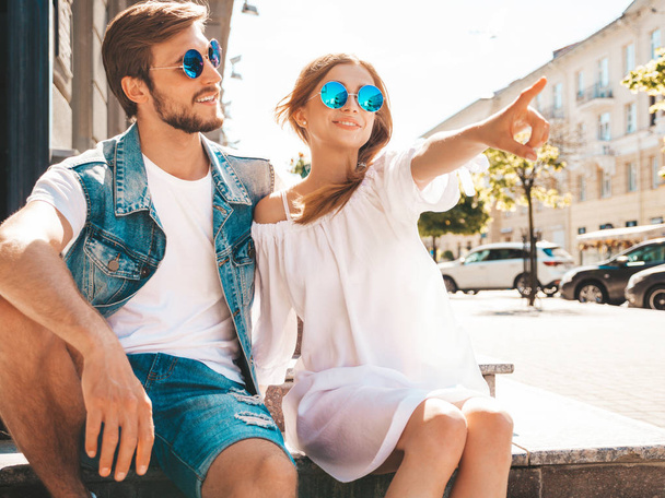 Smiling beautiful girl and her handsome boyfriend.Woman in casual summer dress and man in jeans clothes. Happy cheerful family.Sitting on stairs in street.Points on something interesting.In sunglasses - Foto, imagen