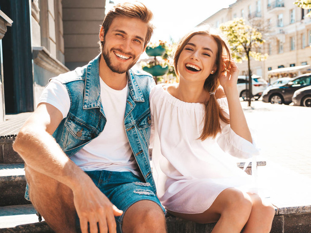 Smiling beautiful girl and her handsome boyfriend. Woman in casual summer dress and man in jeans clothes. Happy cheerful family. Sitting on stairs on the street background.Hugging couple - Fotó, kép