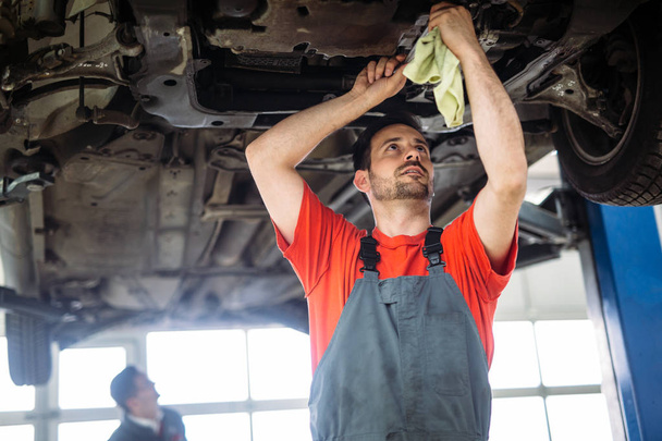 Two handsome mechanics in uniform are working in auto service with lifted vehicle. - Foto, imagen