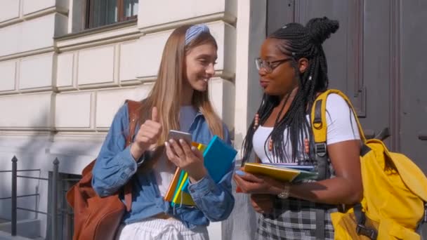 Vooraanzicht van twee mooie studenten van Afrikaanse en Europese afkomst, die samen staan. Vrouwen studeren samen in een luxueuze internationale school en onderhouden vriendelijke relaties. - Video
