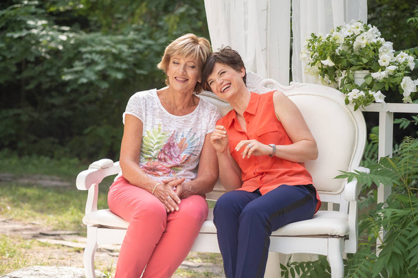 Mother with her adult daughter smiling and hugging have a conversation on the bench outdoor - 写真・画像