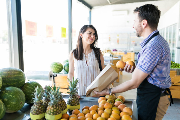 Чоловік власник продуктового магазину упаковує апельсини в папері, розмовляючи з жіночим клієнтом на фруктовій стоянці на органічному ринку
 - Фото, зображення