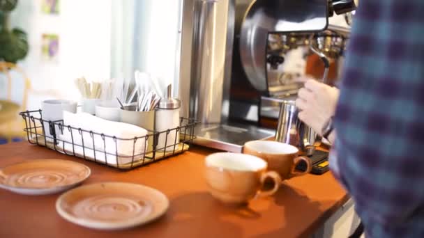 Vista lateral de un barista en camisa a cuadros azul prepara capuchino o latte en su coffeeshop. Art. Poner la leche en la jarra de metal para la preparación del café en la cafetería
. - Metraje, vídeo