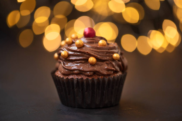 Chocolate cupcake with golden candies on dark wooden background against blurred lights. Selective focus. Unhealthy food. - Valokuva, kuva
