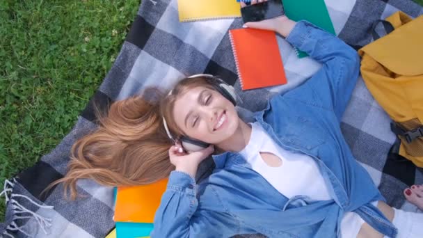 Smile girl student listening music outdoor, lying on grass and relaxing - Materiaali, video