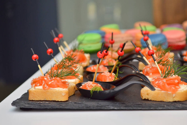 Canapes of bread and red fish, lie on a black stone board on the table at banquet, wooden and glass background - Photo, Image