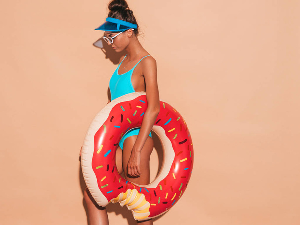 Young beautiful sexy smiling hipster woman in sunglasses.Girl in summer swimwear bathing suit with donut lilo inflatable mattress.Positive female going crazy.Near beige wall in transparent visor cap - Photo, Image