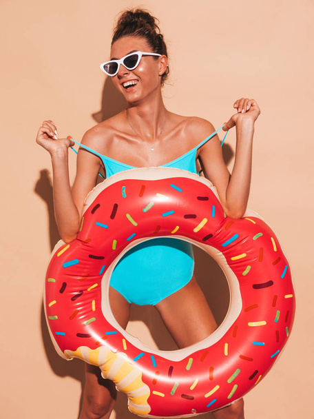 Young beautiful sexy smiling hipster woman in sunglasses.Girl in summer swimwear bathing suit with donut lilo inflatable mattress.Positive female going crazy.Near beige wall in transparent visor cap - Foto, afbeelding