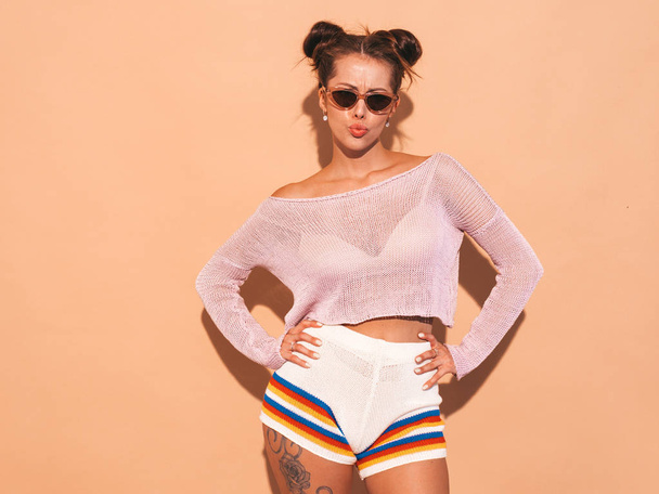 Portrait of young beautiful sexy woman with two horns hairstyle. Trendy girl in casual summer hipster knitted cardigan topic ,shorts in sunglasses. Hot model isolated beige wall.Makes duck face - Photo, image