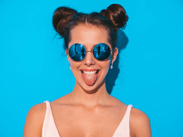 Closeup portrét mladé krasavičky, sexy usměvavé hipíky v slunečních brýlích. Moderní pozitivní holka blázní. Model izolovaný na modré stěně. Zobrazuje jazyk - Fotografie, Obrázek