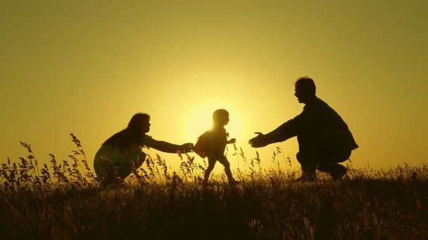 両親と一緒に日没時にジャンプする小さな娘。夜明けの光の中でお母さんと赤ちゃんのシルエット。家族の概念。自然の中で小さな子供と歩く. - 写真・画像