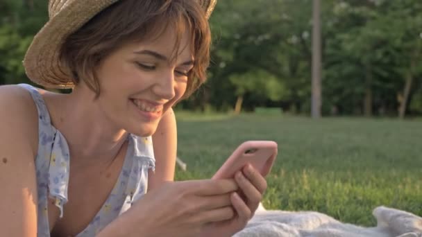 Close up view of Cute smiling young woman chatting on smartphone and laughing while lying on a plaid in the park  - Video