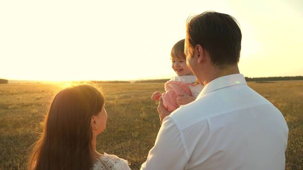 Ο μπαμπάς και η μαμά παίζουν με μια μικρή κόρη στην αγκαλιά της το ηλιοβασίλεμα. οικογενειακές βόλτες με ένα παιδί κατά τη δύση του ηλίου. πατέρας να παίζει με τις κόρες του να ξεκουράζονται στο πάρκο. έννοια της ευτυχης οικογένειας και της παιδικής ηλικίας. - Φωτογραφία, εικόνα