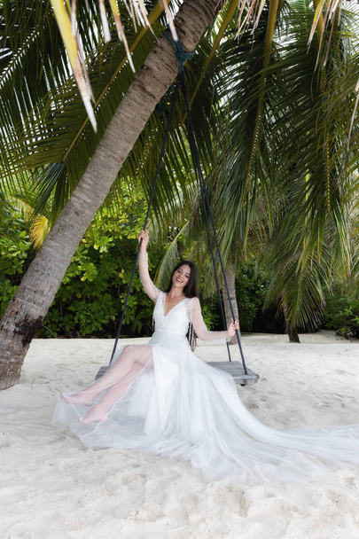 A bride in a white dress is riding on a swing under a big palm tree. - Valokuva, kuva