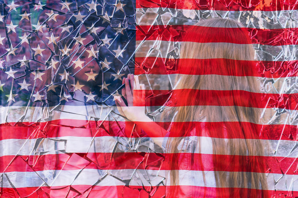 junges Mädchen blickt in einen zerbrochenen Spiegel und zeigt ihre Hand auf einen Spiegel vor dem Hintergrund der amerikanischen Flagge. zieht seine Hand zu seinem Spiegelbild - Foto, Bild