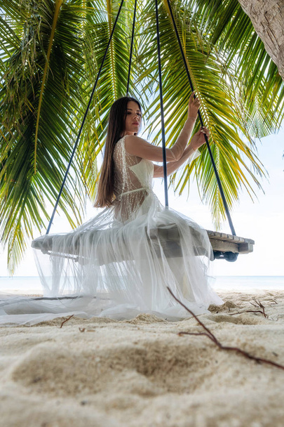 Een bruid in een witte jurk rijdt op een schommel onder een grote palmboom. - Foto, afbeelding