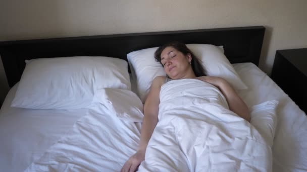 Μελαχρινή νεαρή γυναίκα ξυπνάει στο κρεβάτι με λευκά σεντόνια. - Πλάνα, βίντεο