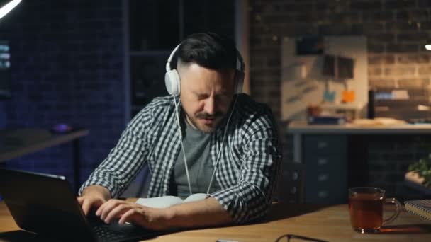 Cheerful man in headphones dancing in office at night having fun then working - Video, Çekim