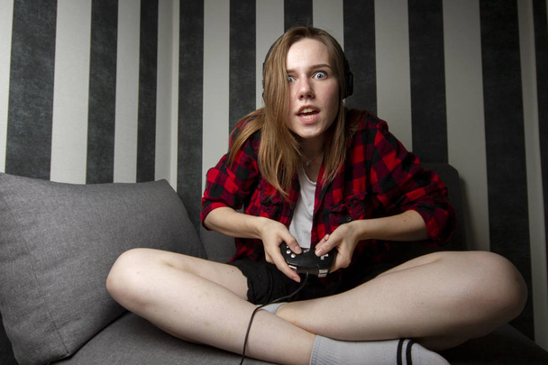 若い女の子はソファに座って、ジョイスティックのコンソールを再生し、彼女はビデオゲームに焦点を当てています - 写真・画像