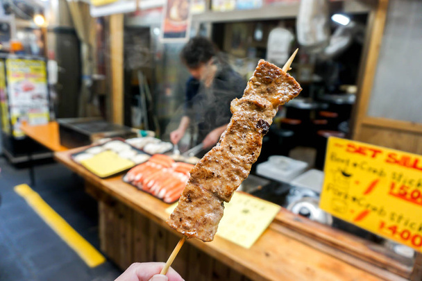 Closeup hand of tourist holding and eat barbecue Kobe beef wood stick on blurry barbecue Kobe beef shop at Kuromon market, Osaka, Japan. - Photo, Image