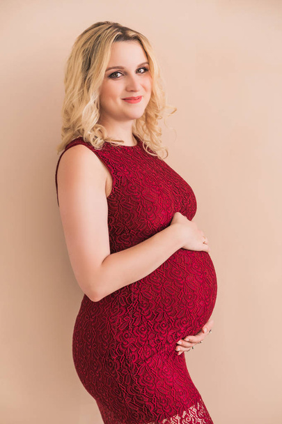 pregnant woman wearing a burgundy dress hugging her belly against a peach-colored wall - Photo, Image