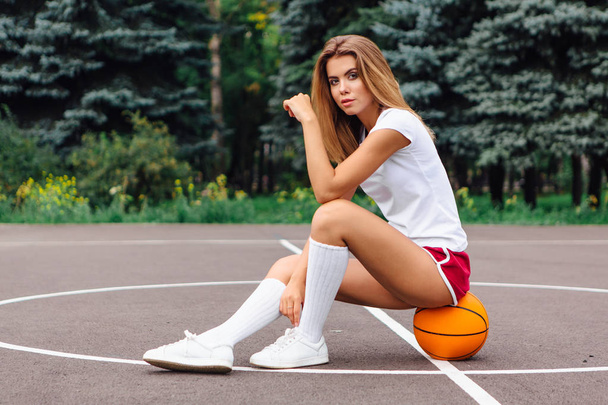 Beautiful young girl dressed in white t-shirt, shorts and sneakers, sits on a basketball court on the ball. - Zdjęcie, obraz