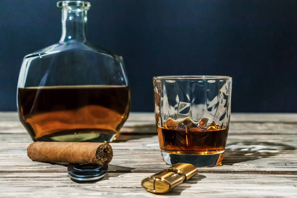Foto en contraste de un vaso de whisky, un cigarro, un encendedor con una guillotina y una botella de alcohol están en la vieja mesa de madera texturizada. Tras un fondo azul oscuro
. - Foto, imagen