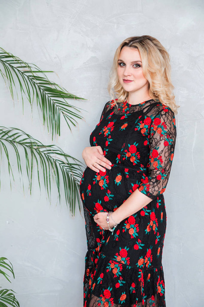 pregnant woman in a black dress is standing against the gray wall and hugging her belly - Fotografie, Obrázek