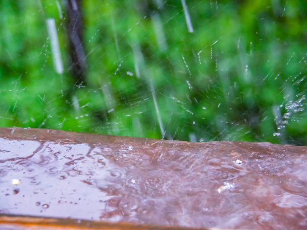 Lluvia fotografiada desde una ventana abierta, vista cercana. Gotas de lluvia en la cornisa marrón. Enfoque suave selectivo. Valla de hierro, hierba verde y hojas, fondo abstracto borroso
. - Foto, Imagen