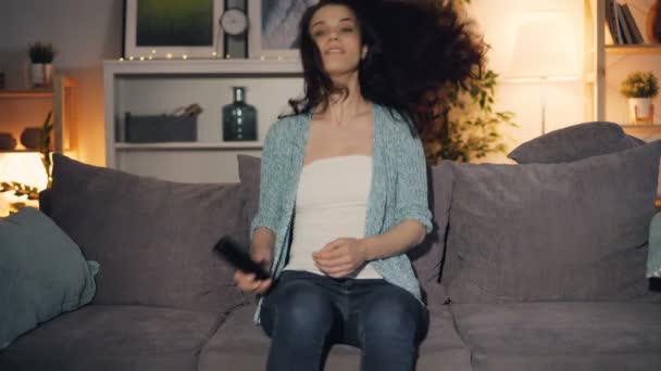 Cute girl turning on TV with remote control watching sitting on sofa at home - Záběry, video