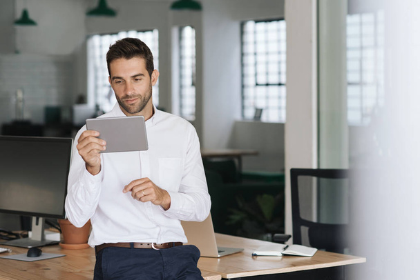 konzentrierter junger Geschäftsmann lehnt an einem Tisch in einem großen modernen Büro und arbeitet online mit einem digitalen Tablet - Foto, Bild