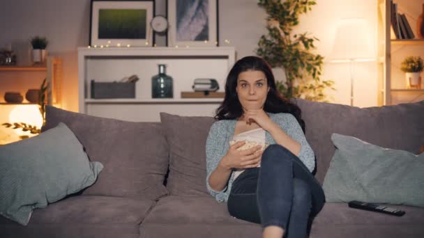 Young lady watching TV and eating popcorn at home sitting on couch alone - Filmagem, Vídeo