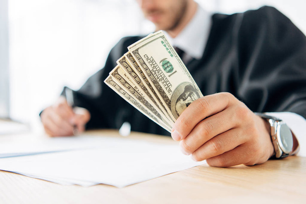 selective focus of judge holding dollar banknotes and writing in office  - Photo, Image
