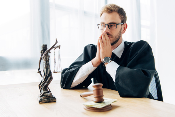 selective focus of judge in glasses covering face near wooden gavel with cash and statuette of justice  - Photo, Image